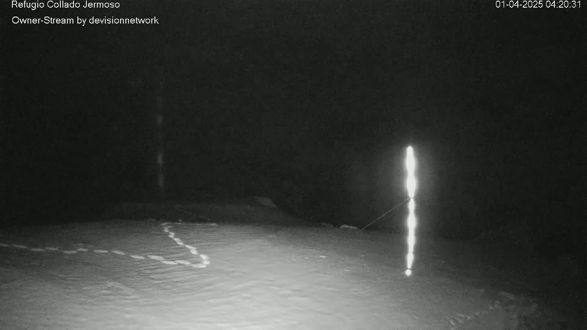 A snow-covered ground at night is illuminated by a vertical line of light, with faint animal tracks visible in the snow.