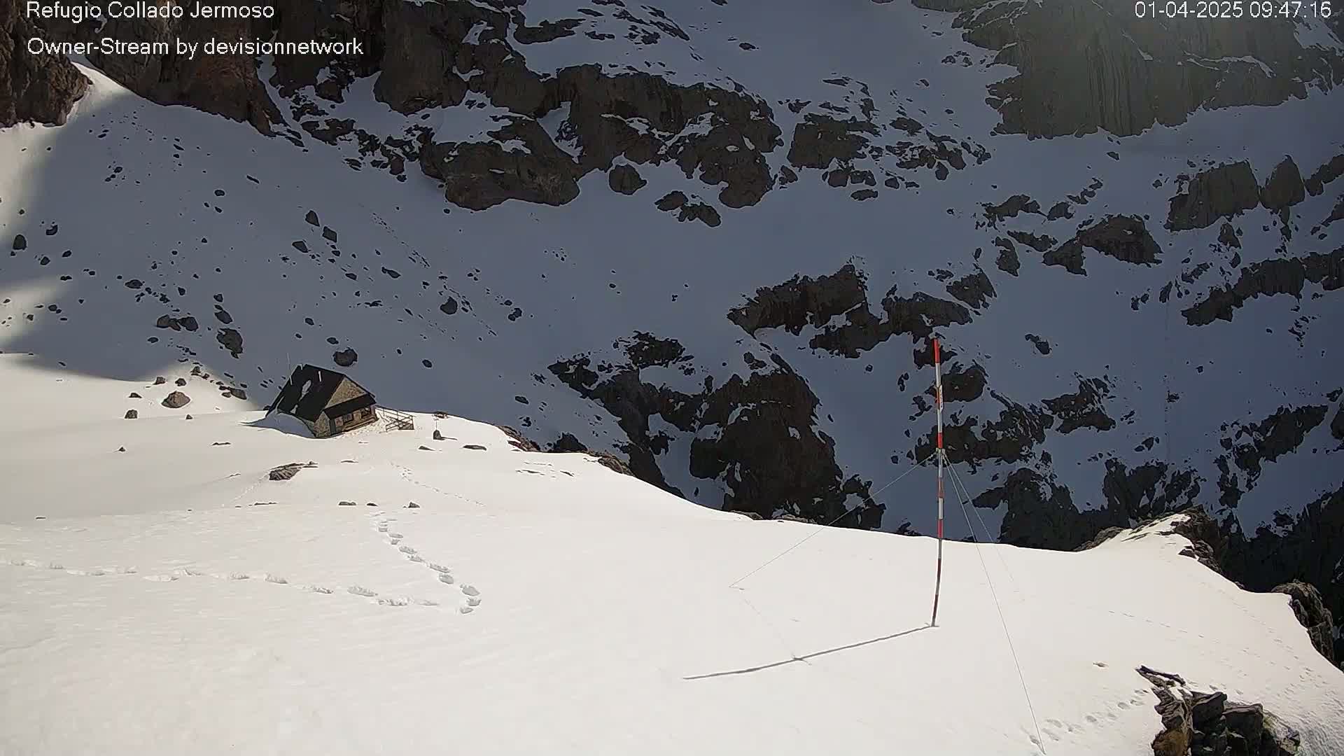 A small cabin sits on a snow-covered mountain peak under sunny conditions, with a tall pole and wires extending towards a deep snowy ravine.