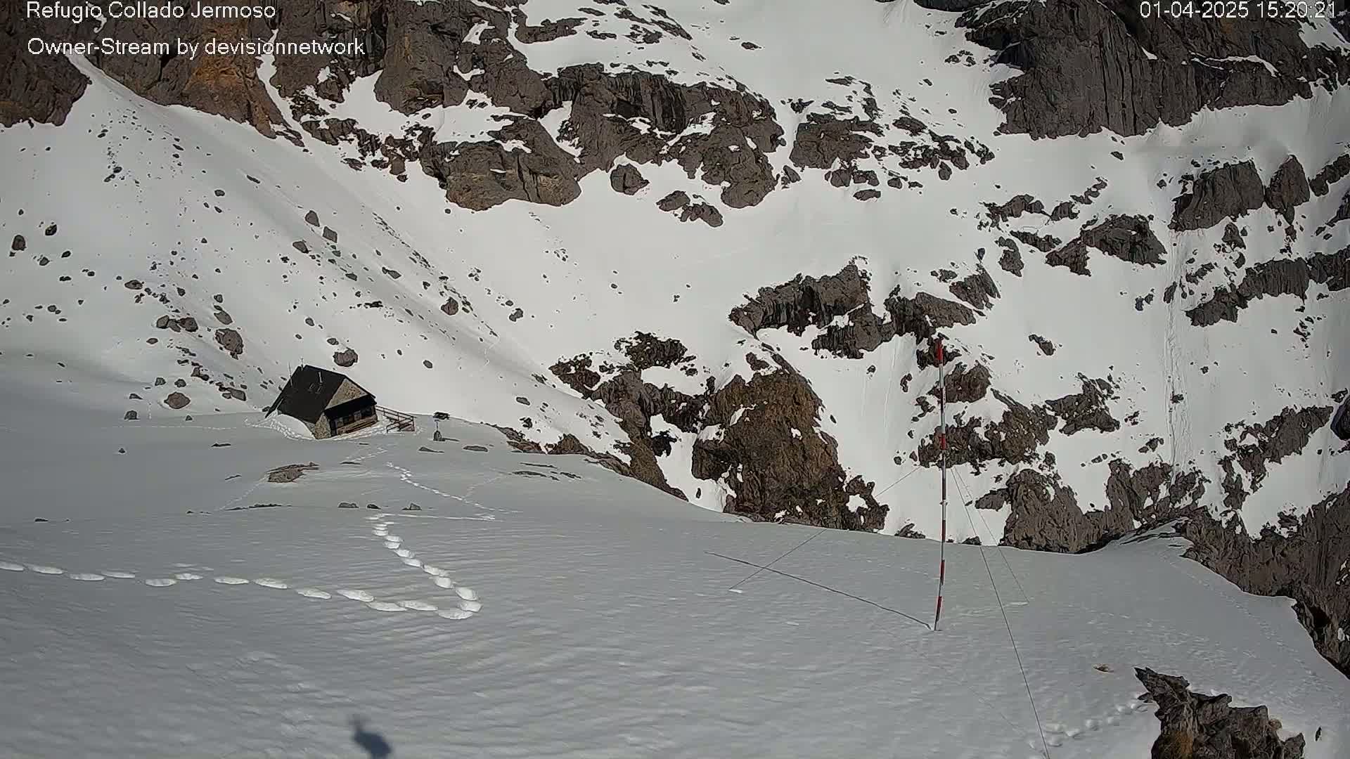 A small, dark building sits on a snow-covered mountainside, with footprints leading away from it and a tall, red-and-white pole nearby, under a bright, sunny sky.