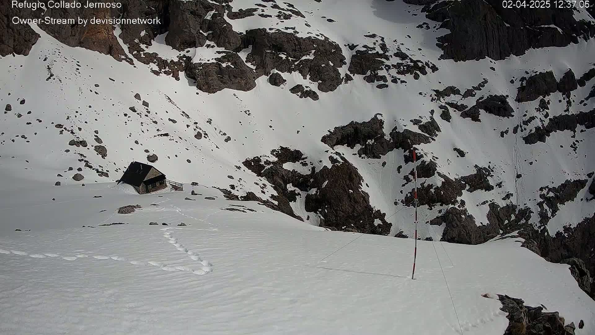 A small, dark building sits nestled in a snow-covered mountain pass, with footprints leading towards it.