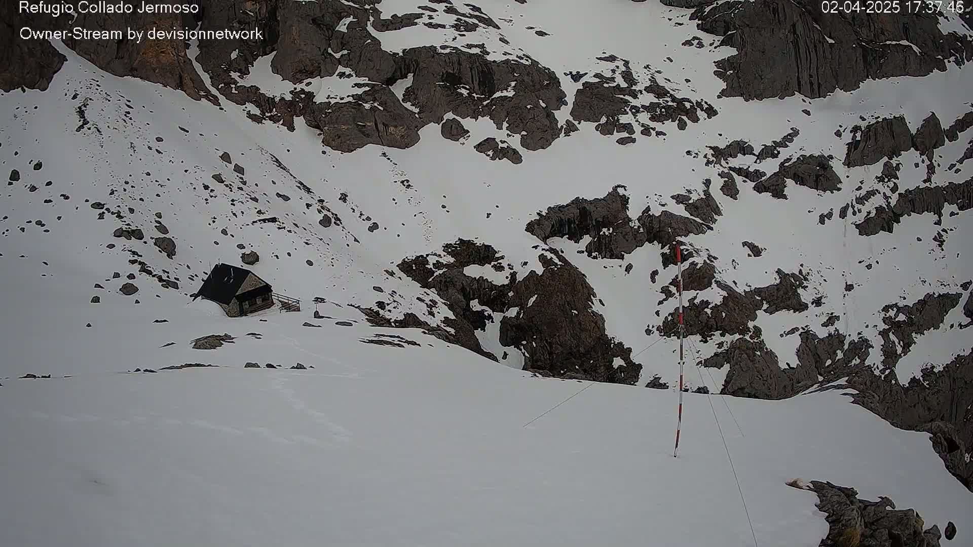 A small cabin sits nestled in a snow-covered mountain pass, with dark rocky outcrops visible amidst the snow.