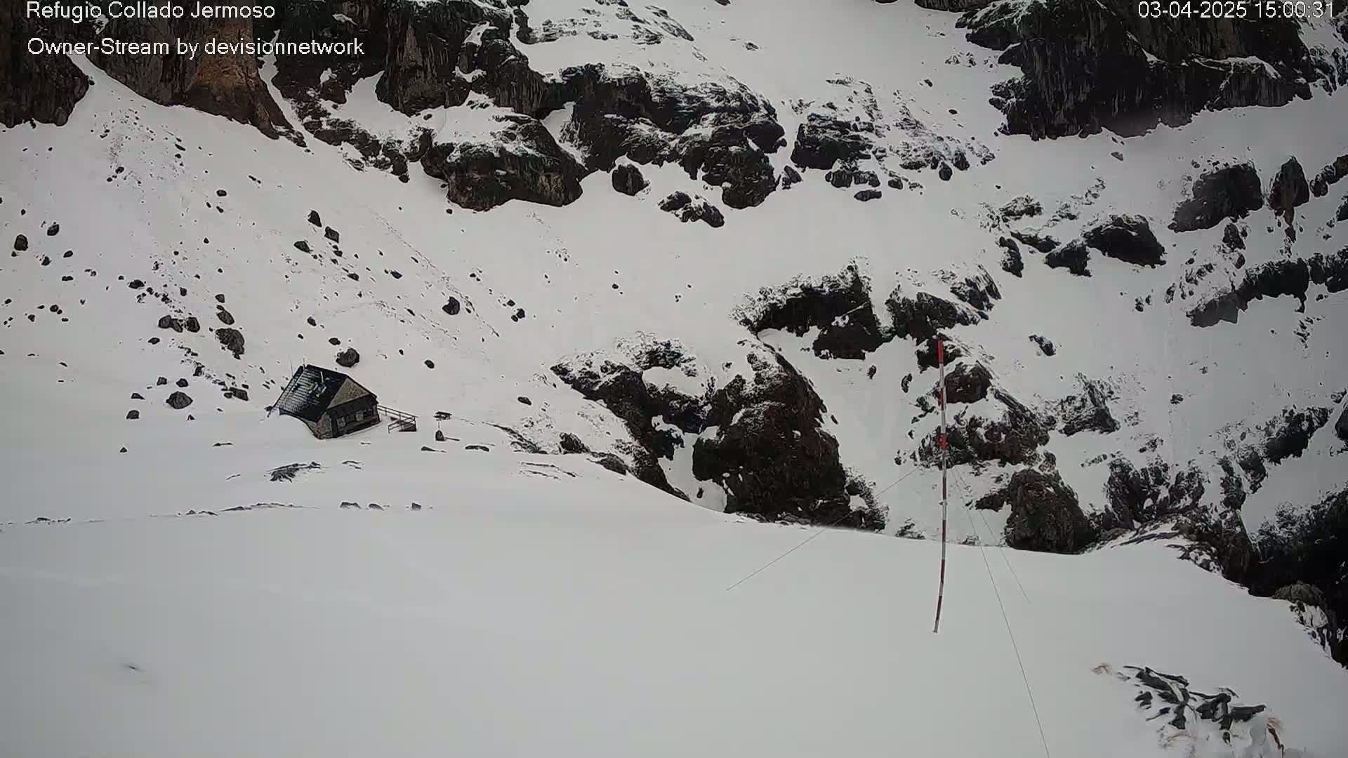A small cabin sits nestled in a snow-covered mountainous area with dark rocks and a tall pole.