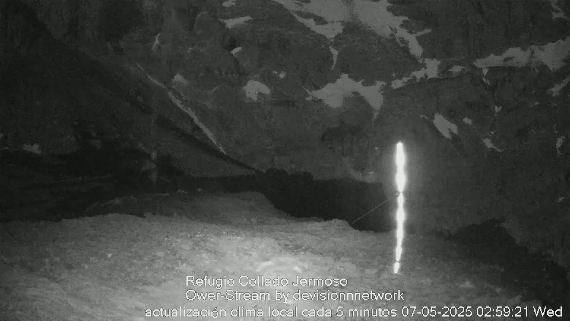 A nighttime, grainy black and white image shows a rocky, mountainous area with a bright vertical light source in the foreground.