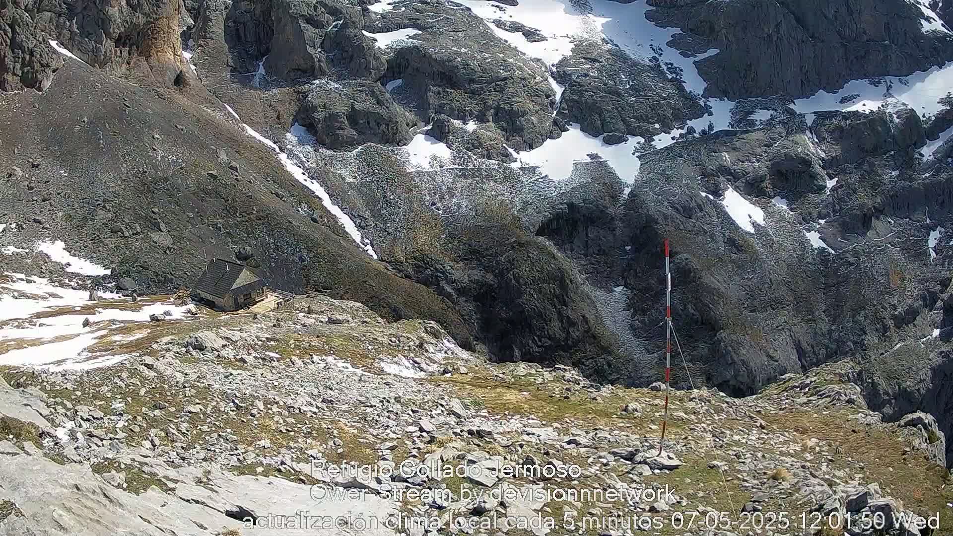 A small cabin sits on a rocky mountainside with patches of snow and grass, under a sunny sky.
