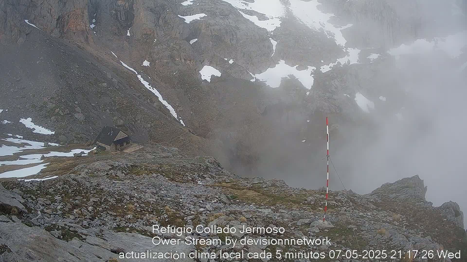 A small stone building sits on a rocky mountainside shrouded in fog, with patches of snow visible on the ground and surrounding peaks.