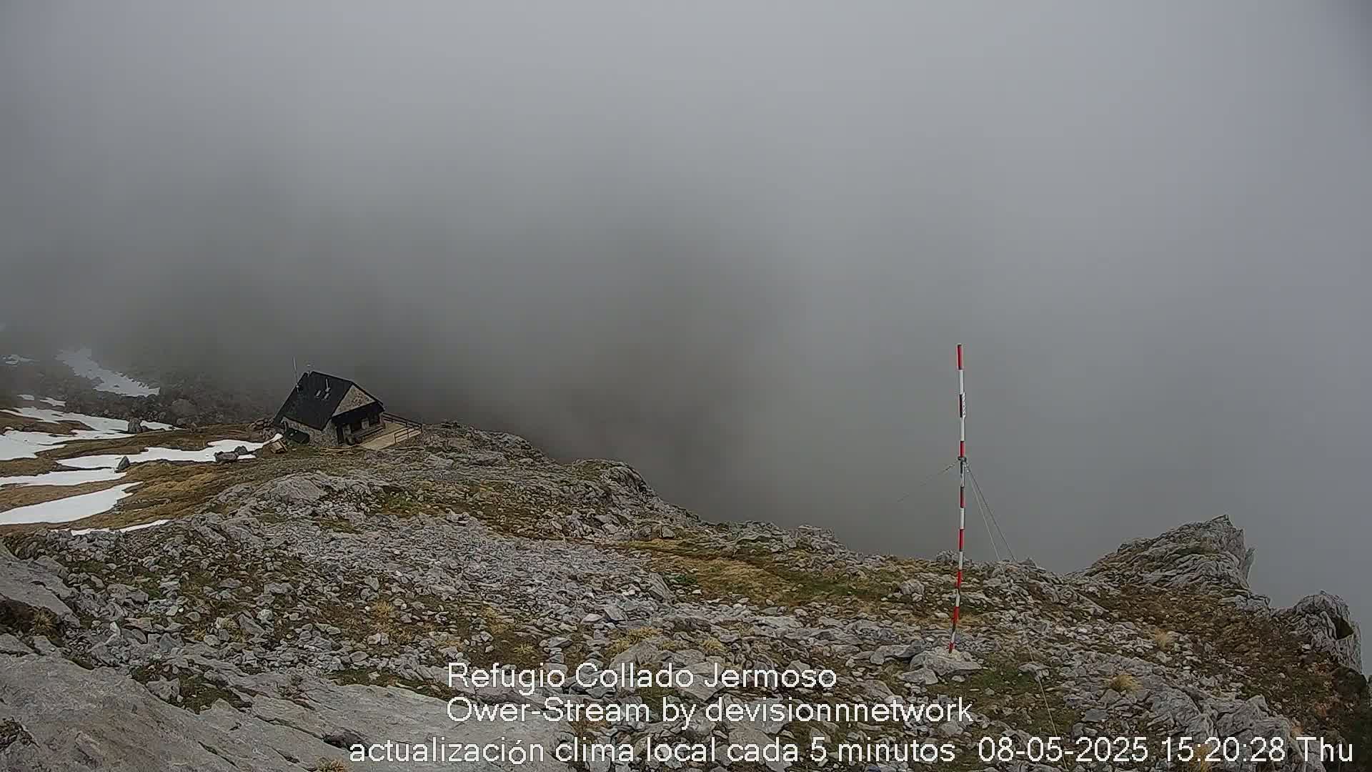A small stone hut sits on a rocky, snow-dusted mountaintop shrouded in thick fog.