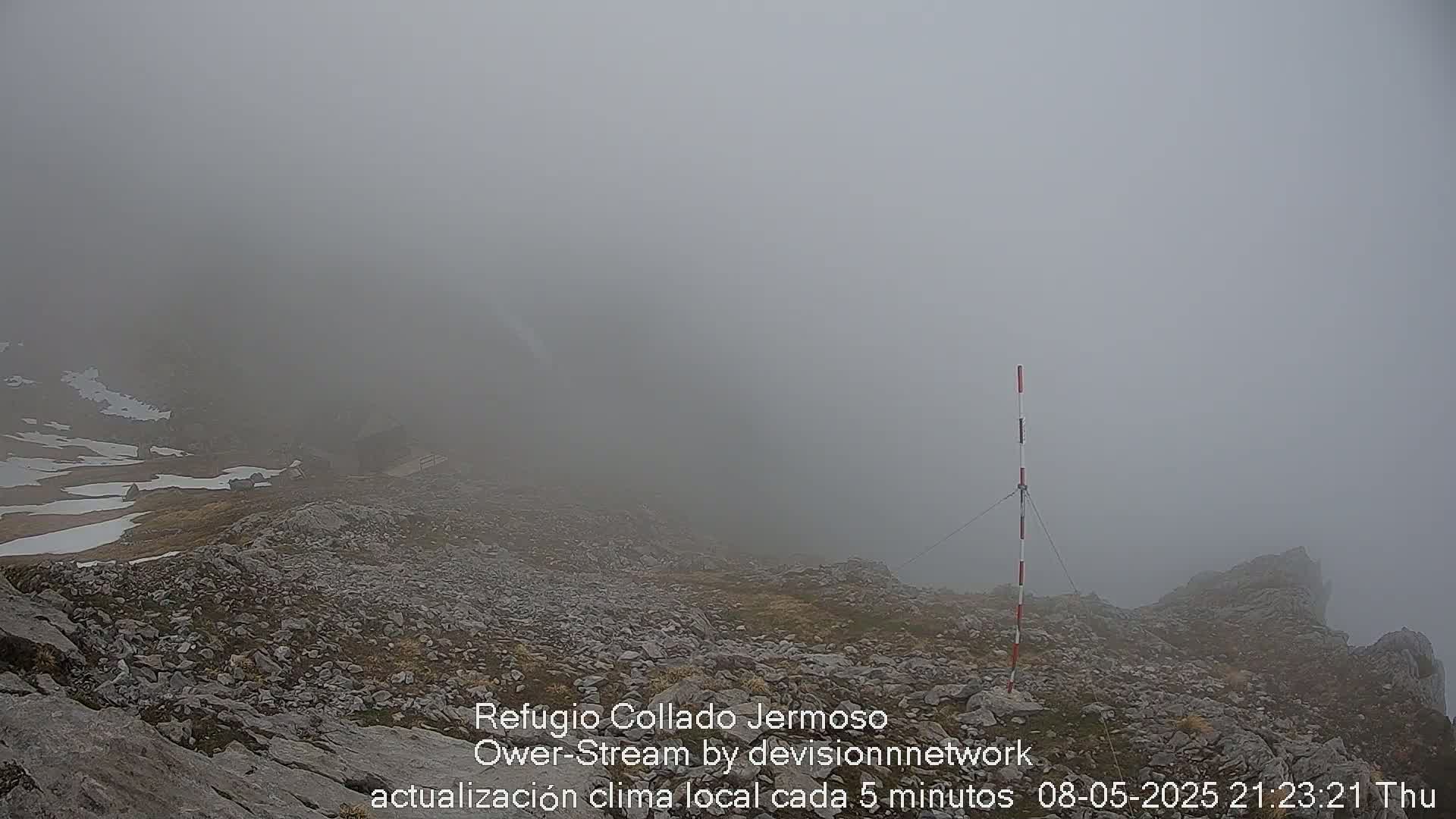 A rocky, fog-shrouded mountaintop scene features a tall, marked pole and patches of snow.