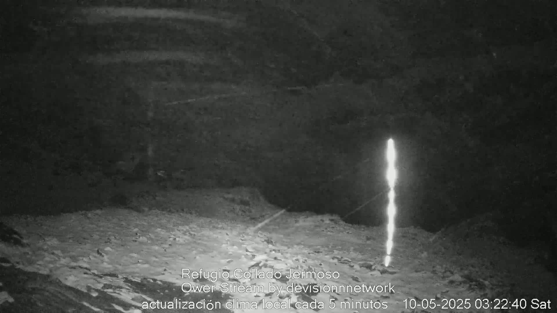 A bright, vertical light source illuminates a dark, rocky, snow-covered ground at night.