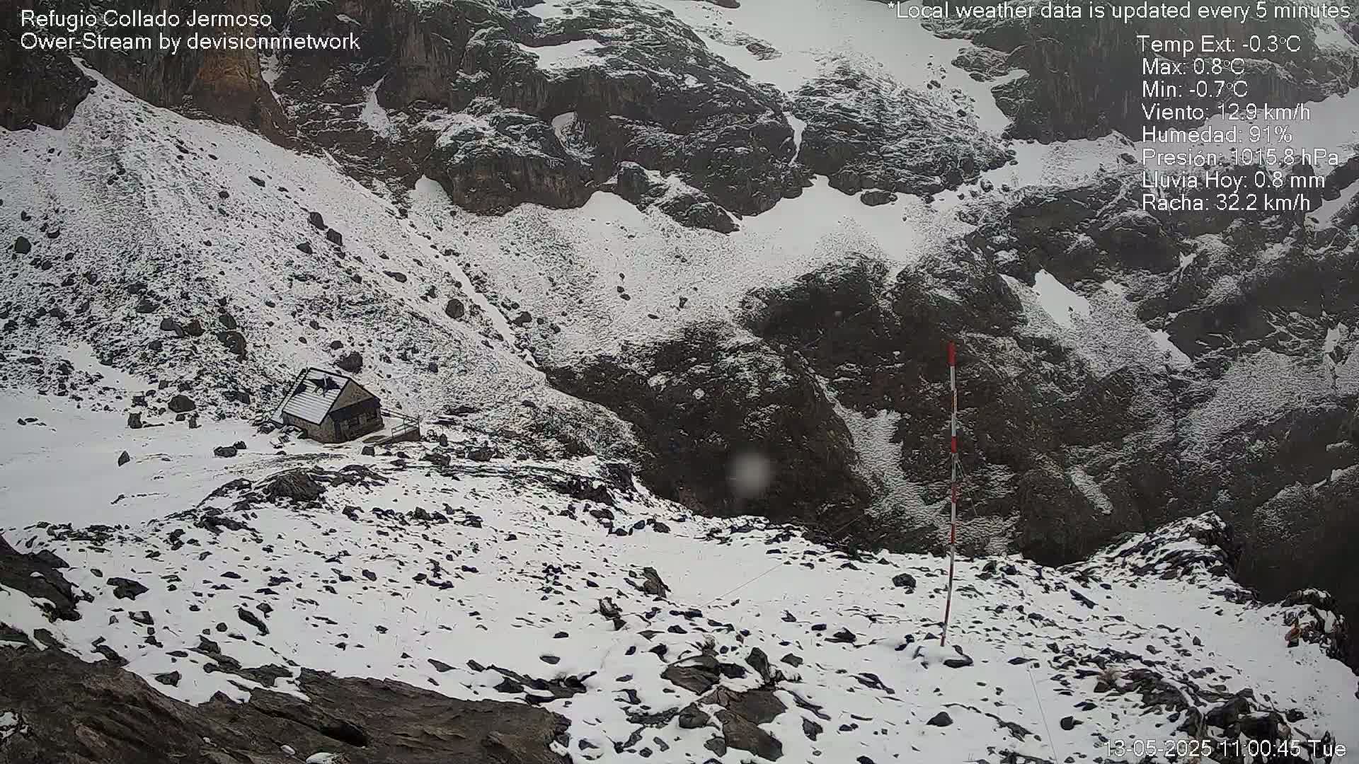 A small stone structure sits on a snow-covered, rocky mountainside in windy conditions.
