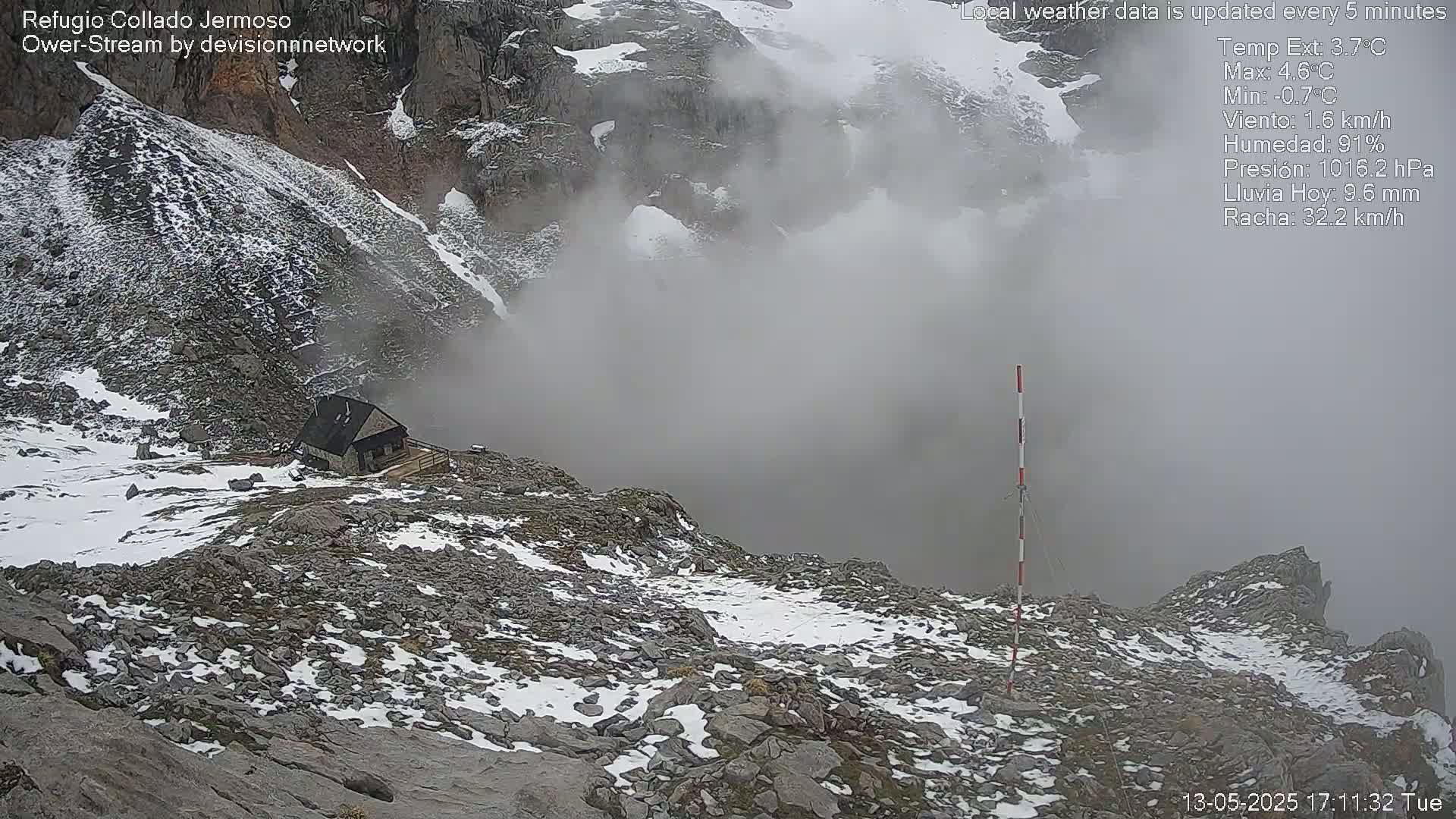 A small stone building sits on a rocky, snow-dusted mountainside shrouded in thick fog.