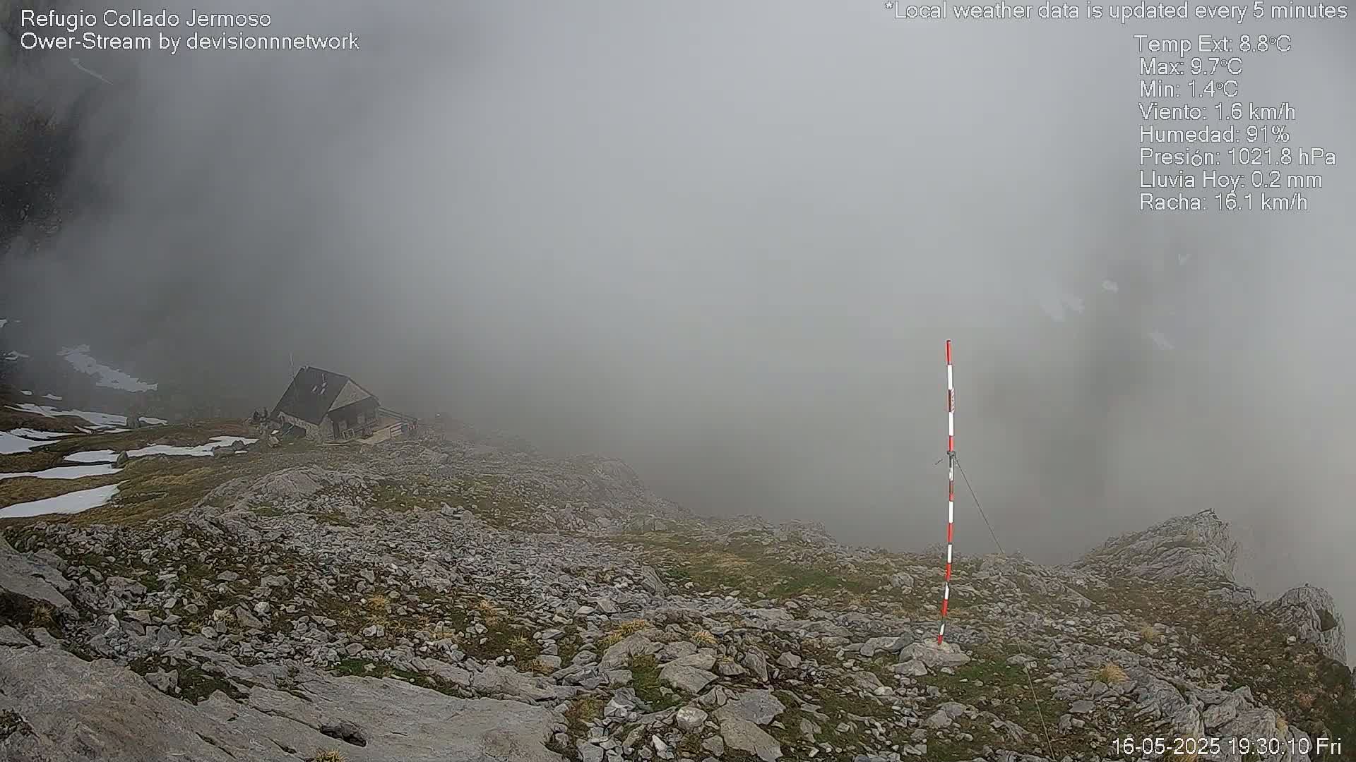 A small cabin sits on a rocky, sparsely vegetated mountainside shrouded in thick fog.