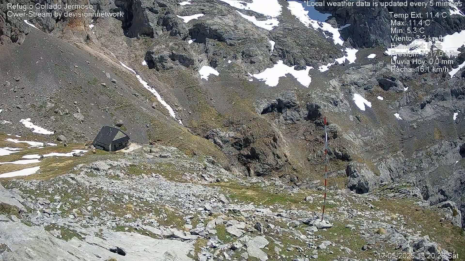 A small stone building sits on a rocky mountainside with patches of snow and sparse vegetation, under a mostly sunny sky.
