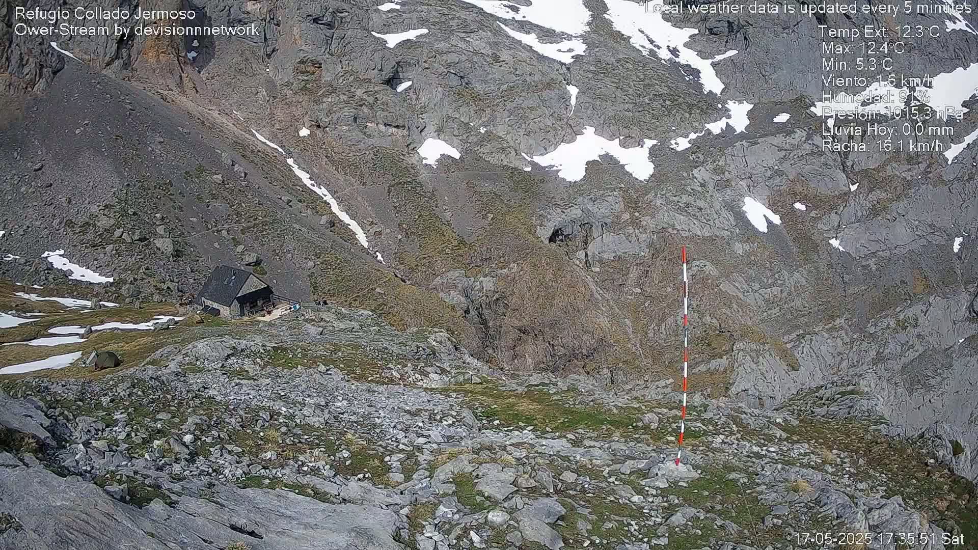 A mountaintop stone hut is visible below a rocky, sparsely vegetated slope with patches of snow, under a partly sunny sky.