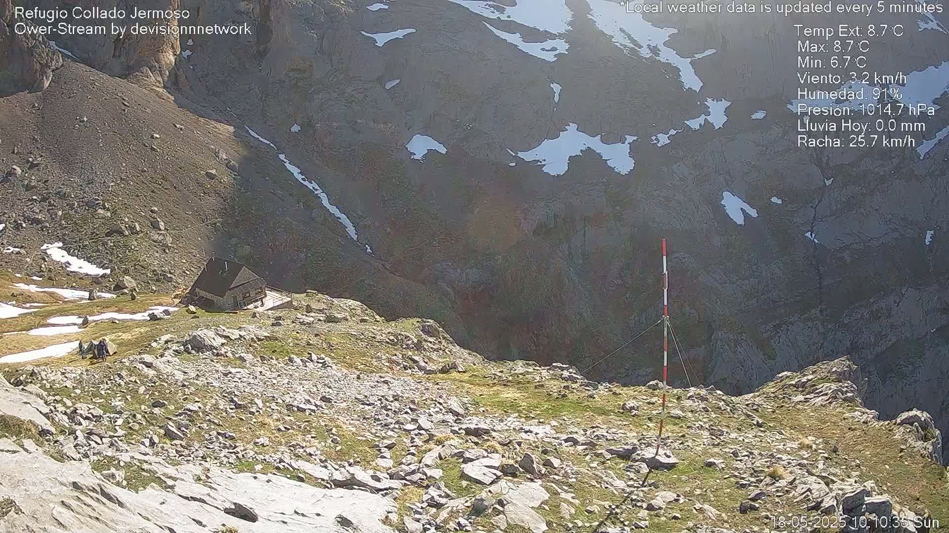 A small cabin sits on a rocky mountainside with patches of snow and sparse vegetation, under sunny skies.