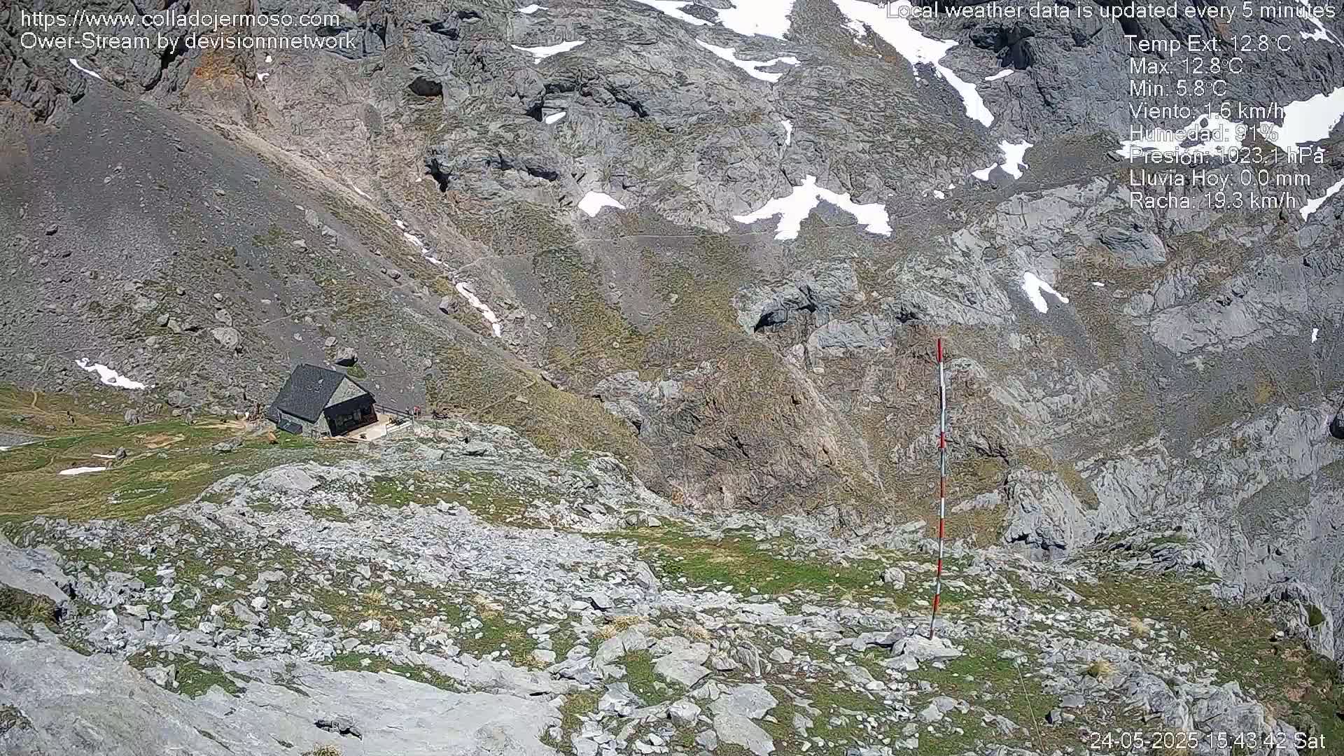 A small building sits on a rocky, mountainous slope with patches of grass and some remaining snow, under sunny conditions.