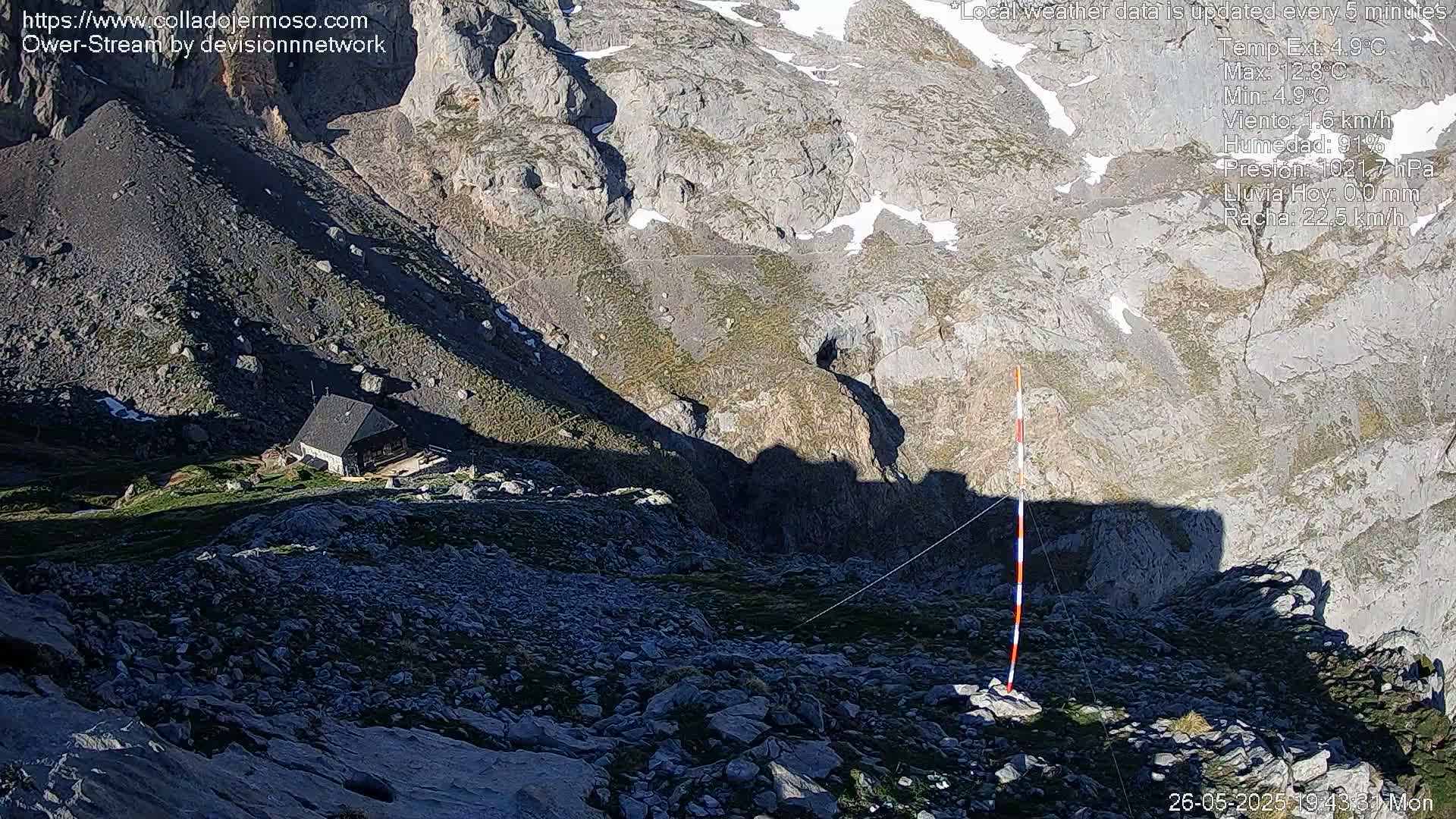 A small building sits on a rocky mountainside partially shaded by shadows, with patches of snow visible in the distance under sunny conditions.