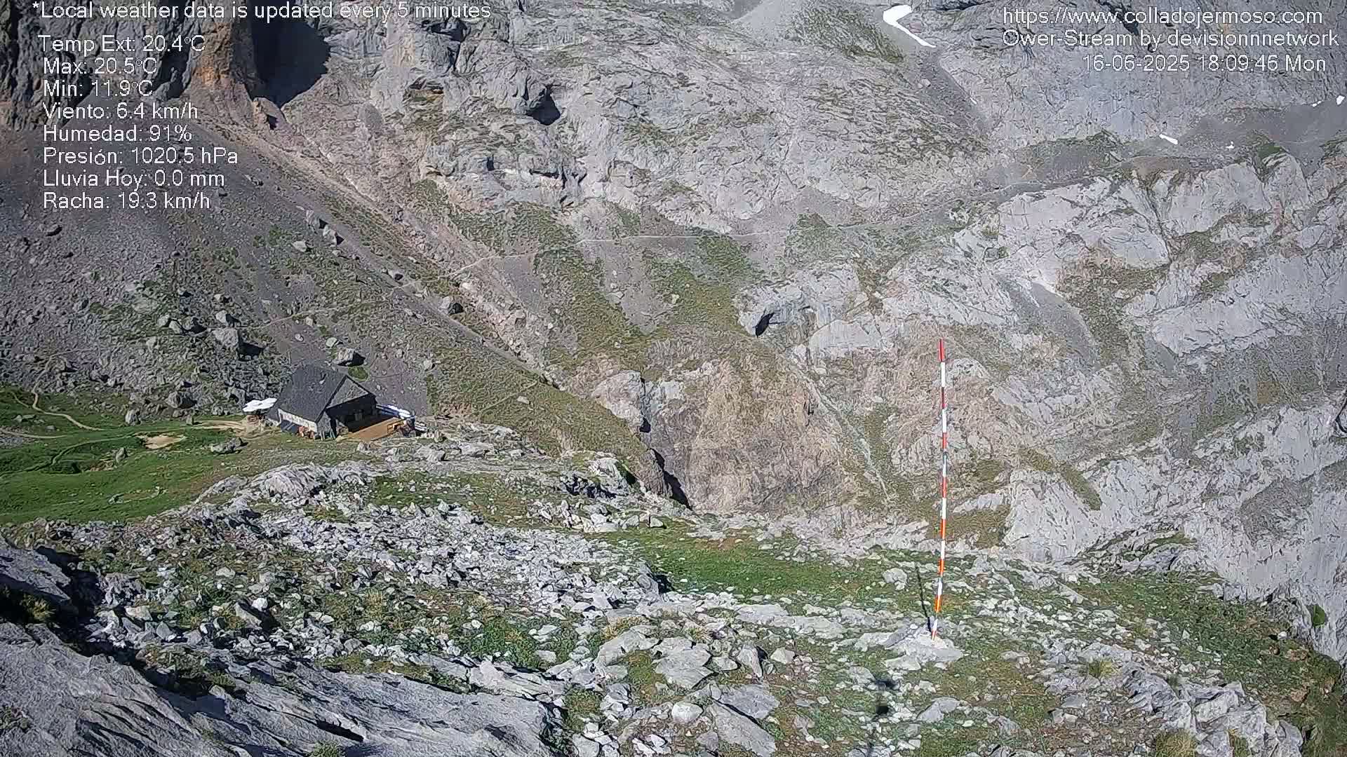 A mountaintop view shows a small building nestled among rocky terrain and sparse vegetation, under sunny conditions.