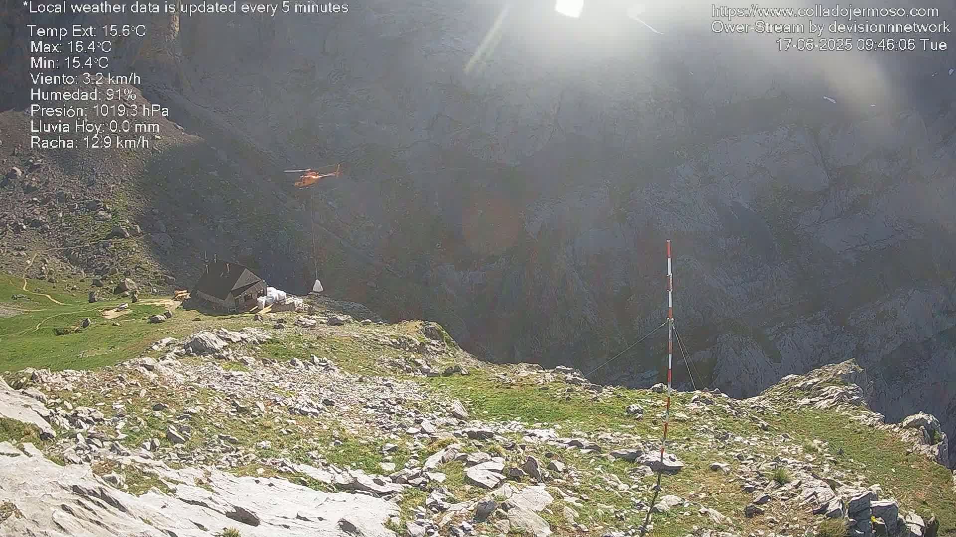 A helicopter flies over a rocky mountainside, near a small building and patches of green vegetation, on a sunny day.