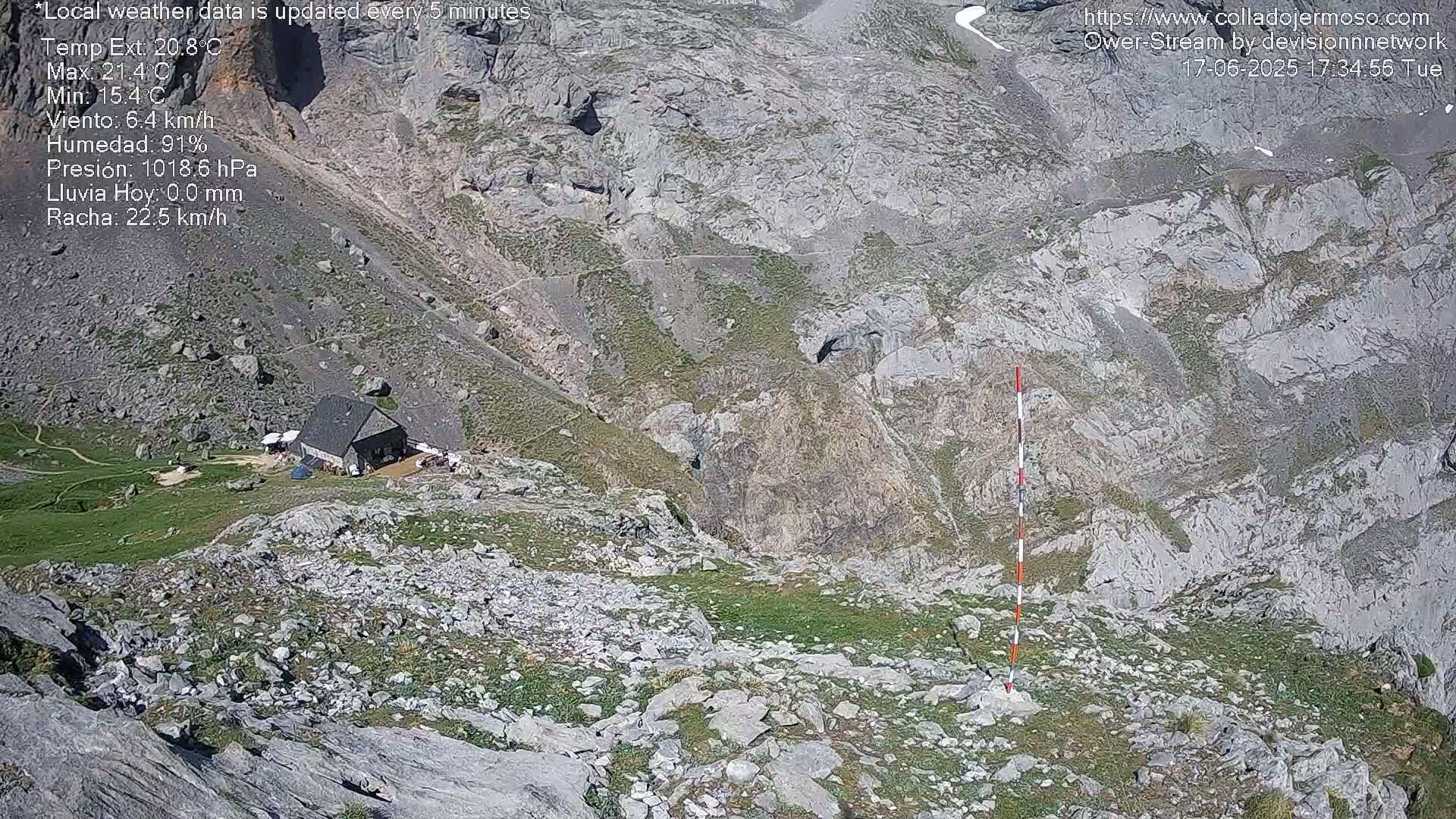 A high-angle view shows a stone building situated in a rocky mountain valley with sparse vegetation under a sunny sky.