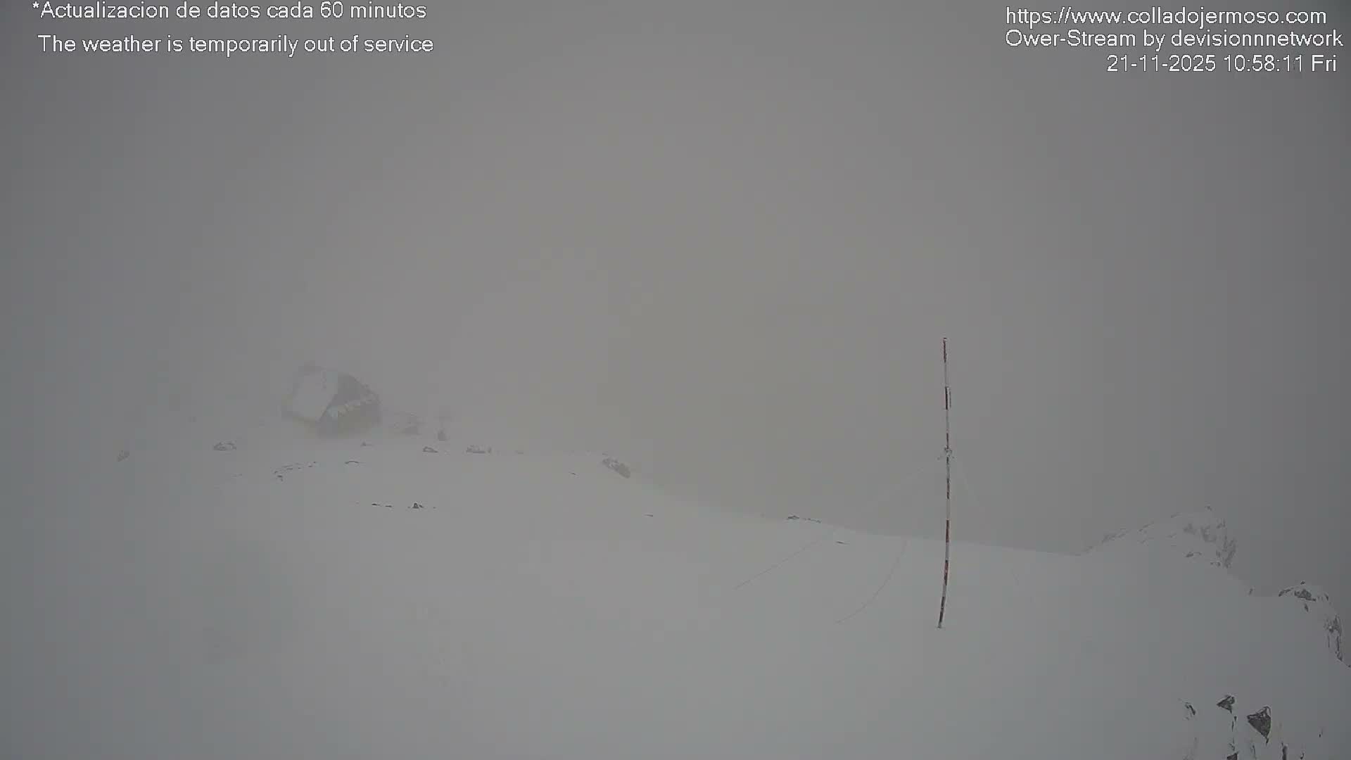 A snow-covered mountain landscape is shrouded in dense fog, with a pole visible on the right and faint rocky outlines in the background under heavily overcast skies.