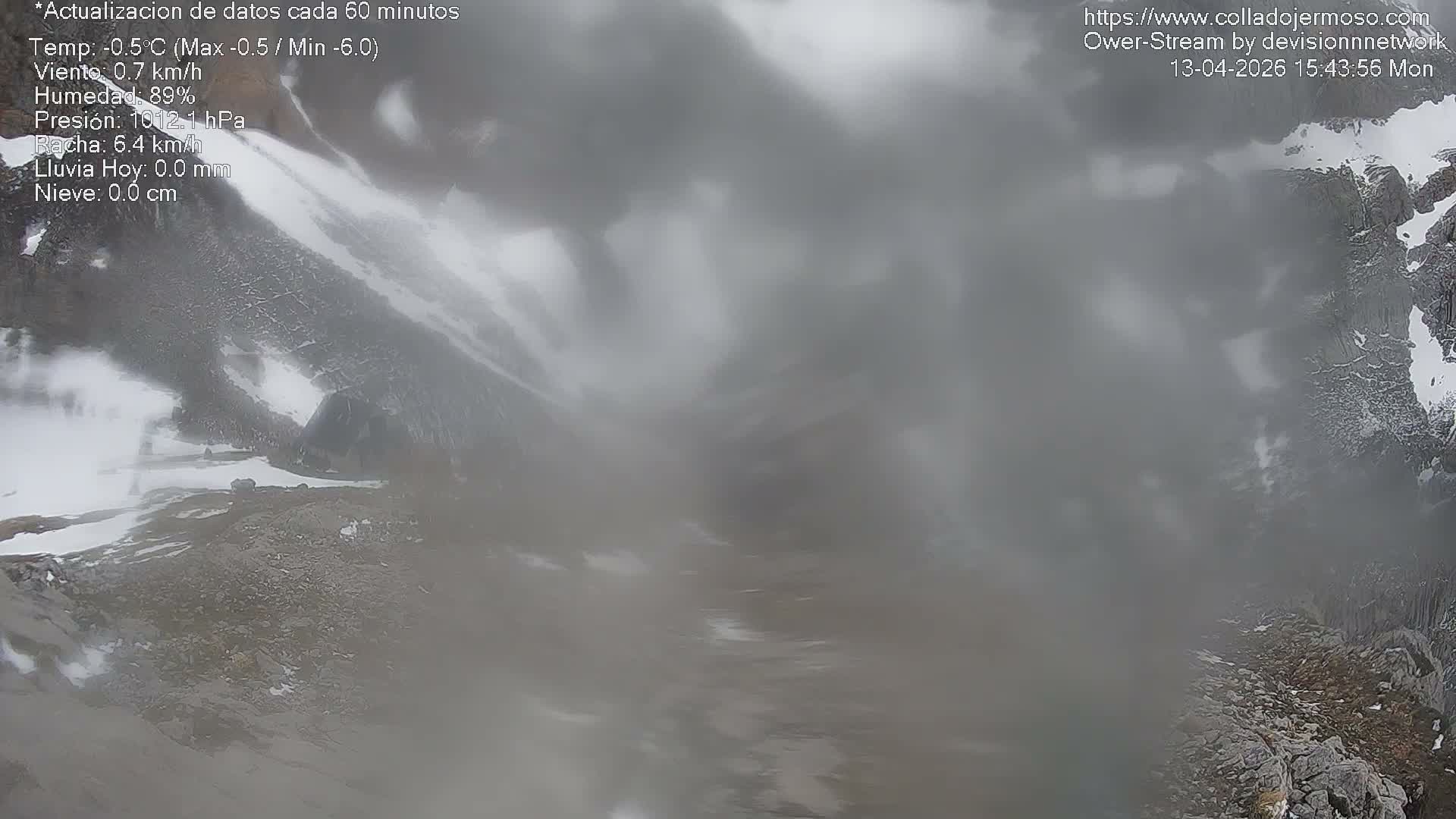 A vast, snow-covered mountain landscape unfolds under an overcast sky, revealing a small building nestled into a snowy slope and a striped marker pole in the foreground, indicating cold, wintry conditions.