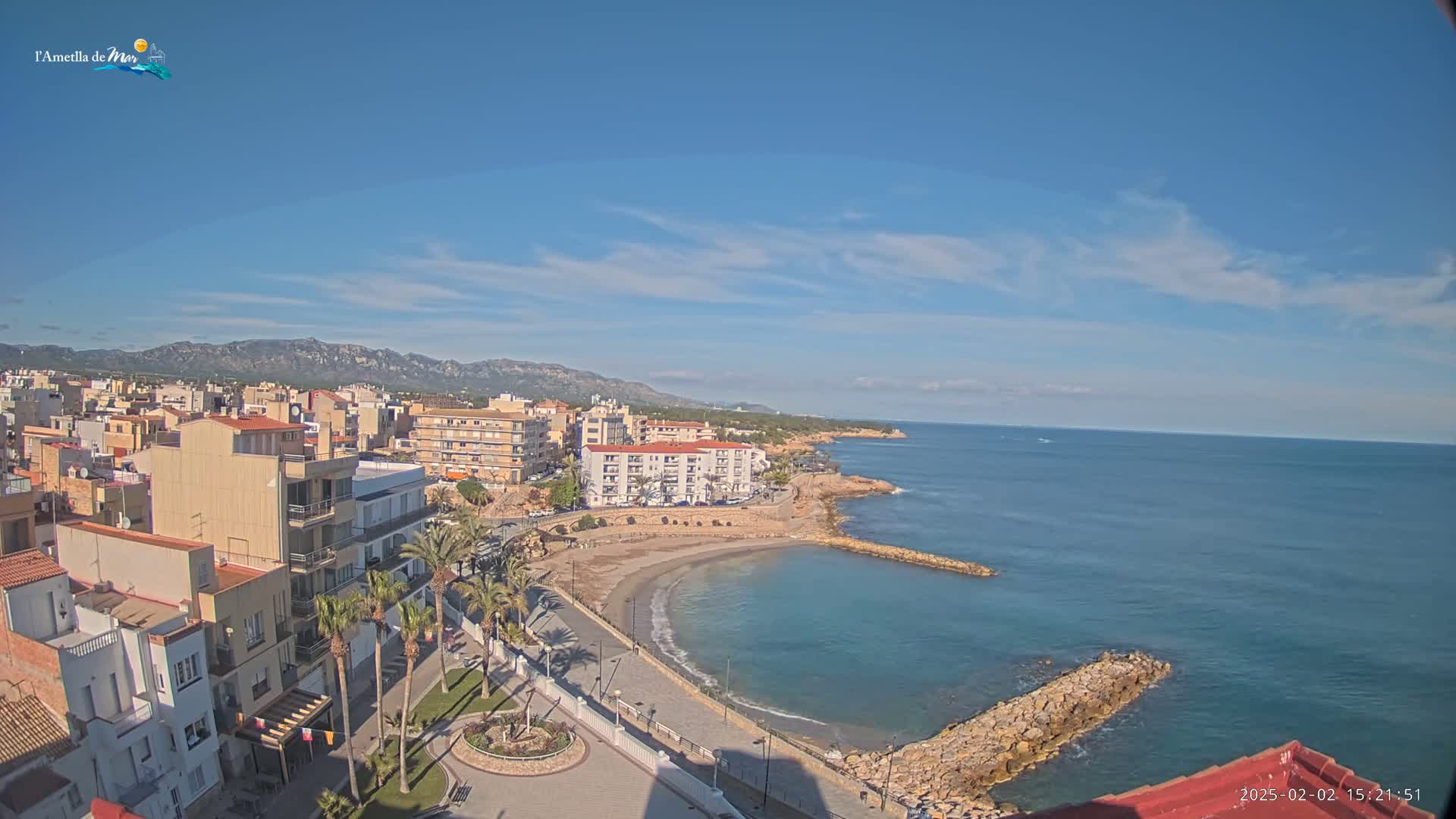 A coastal town with buildings and palm trees sits on a calm, blue sea under a mostly sunny sky with a few scattered clouds and mountains in the distance.