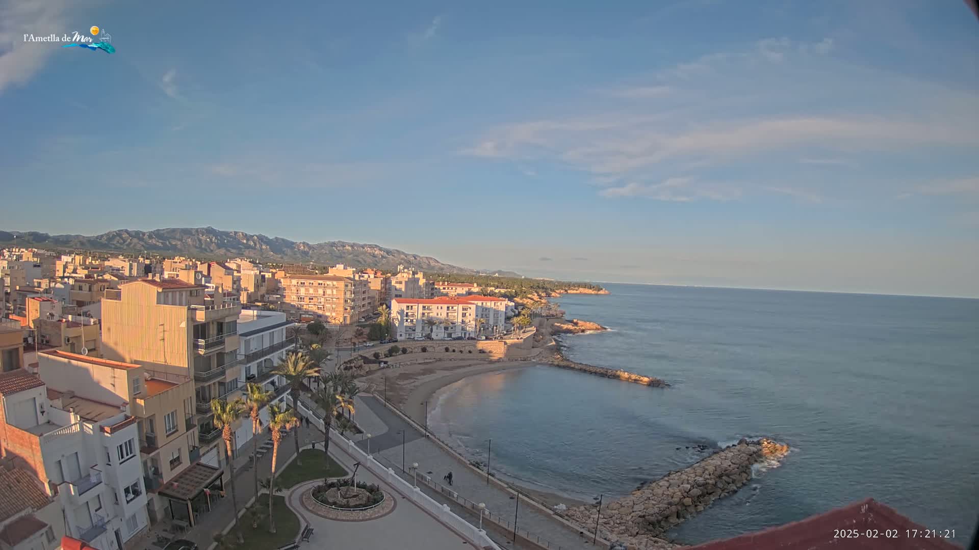 A coastal town with buildings, palm trees, and a small sandy beach is nestled against a calm sea under a mostly clear sky.