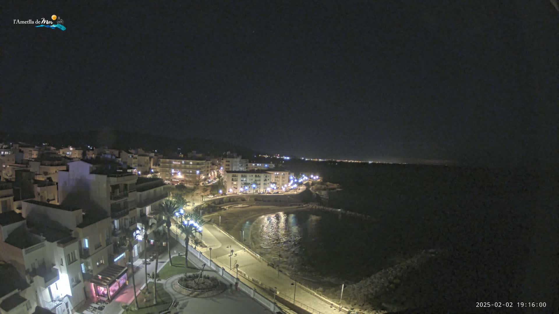 A nighttime, high-angle view shows a coastal town with buildings lit up along a paved waterfront path next to a calm bay under a clear, dark sky.
