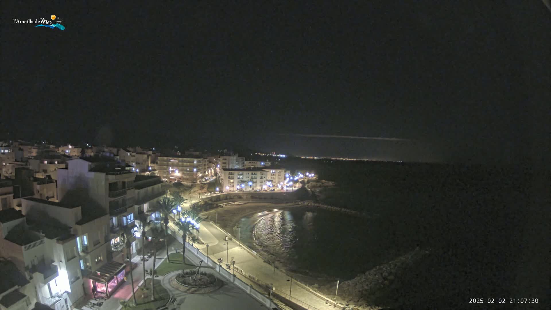 A nighttime view of a coastal town with a small, dark cove,  illuminated buildings lining a paved walkway, and palm trees.