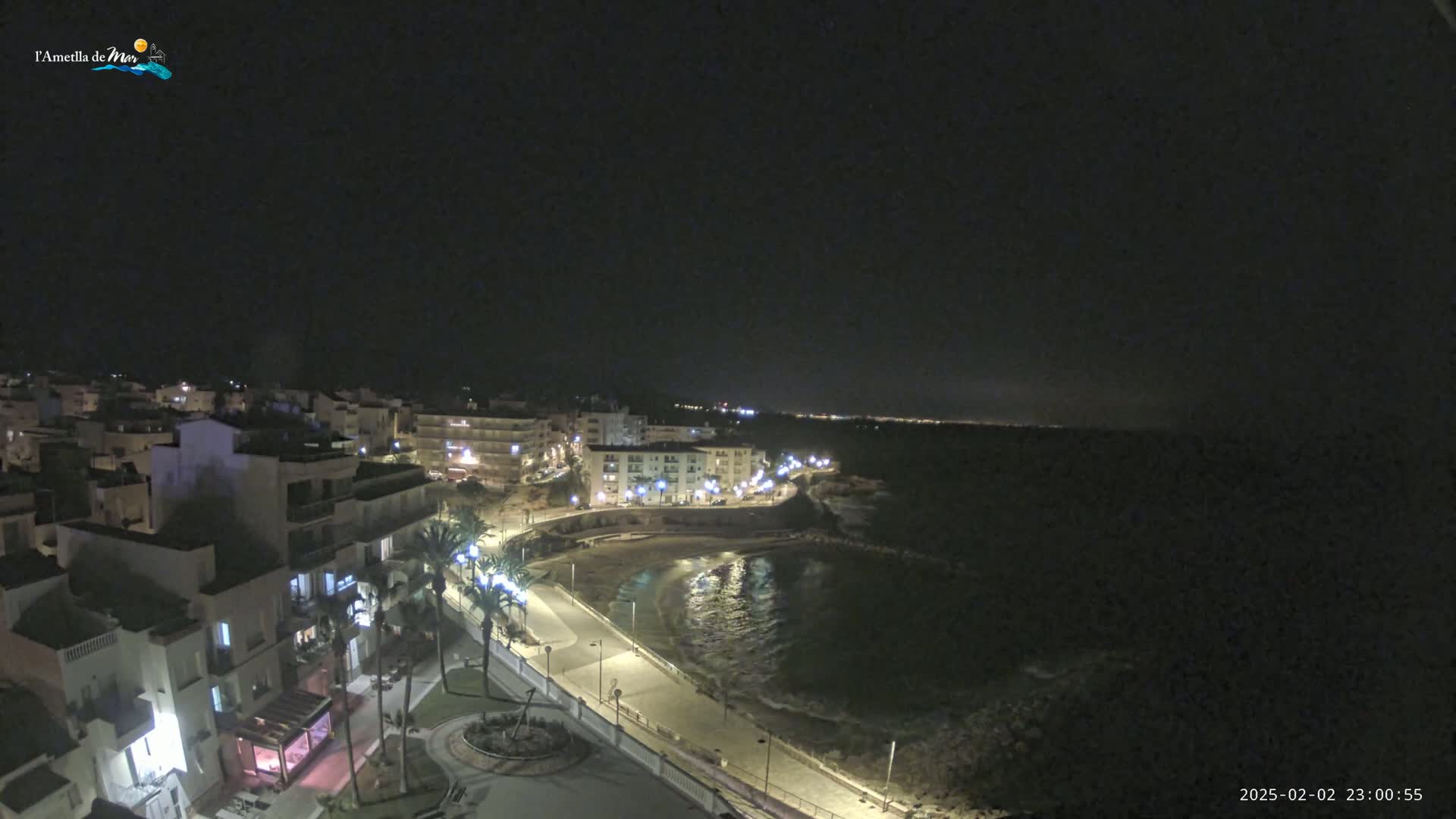 A nighttime, seaside town with a crescent-shaped beach and buildings illuminated along a paved walkway, under a clear, dark sky.