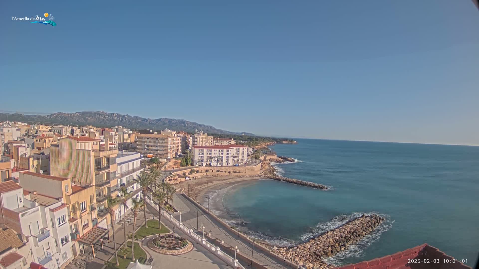 A coastal town with buildings, palm trees, and a small sandy beach is situated next to a calm, blue sea under a clear, sunny sky.