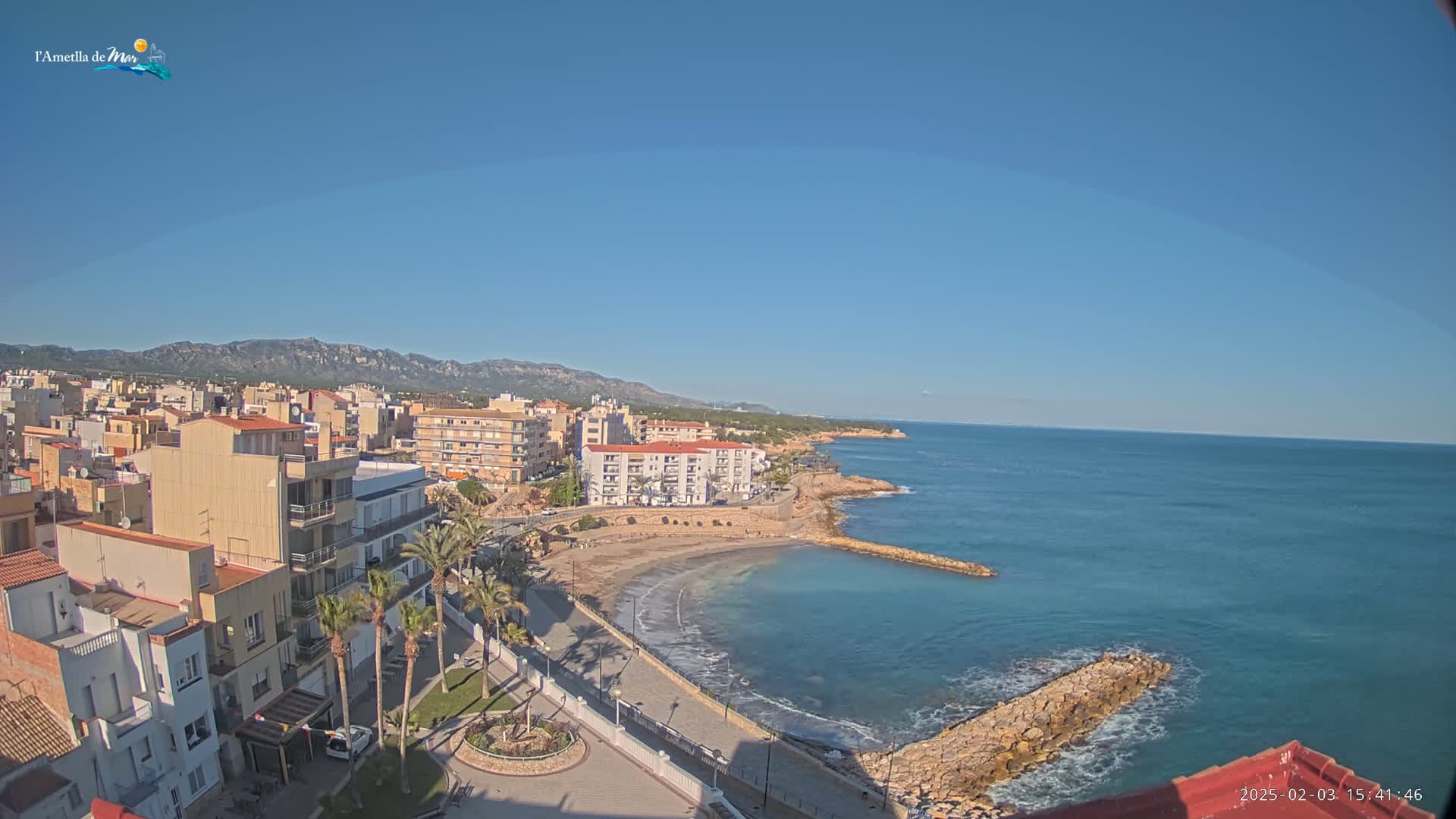A coastal town with buildings, palm trees, and a small sandy beach sits on a clear, sunny day next to a calm sea under a clear blue sky with distant mountains visible in the background.