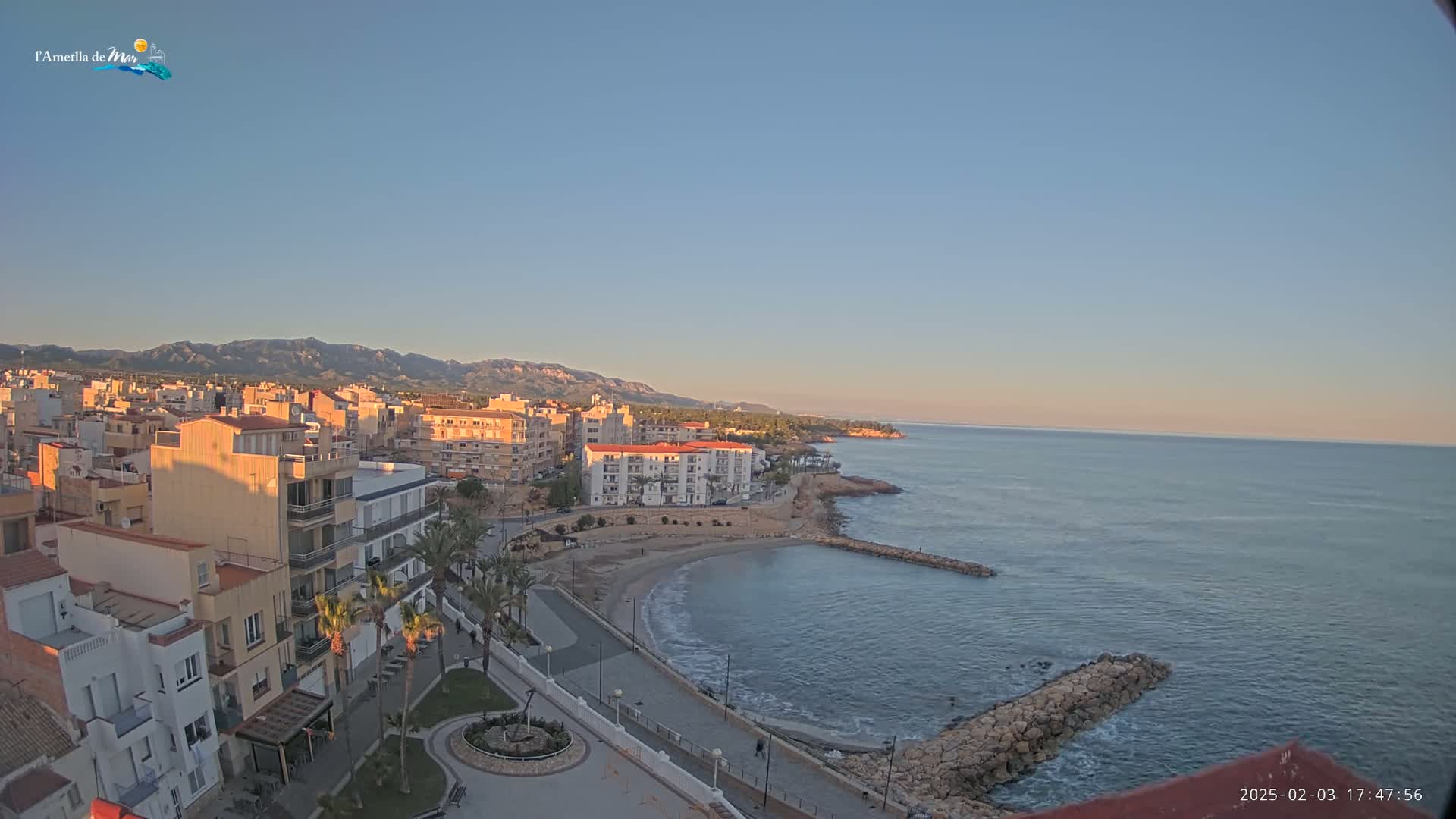 A coastal town with buildings and palm trees sits on a calm sea under a clear, twilight sky, mountains visible in the distance.