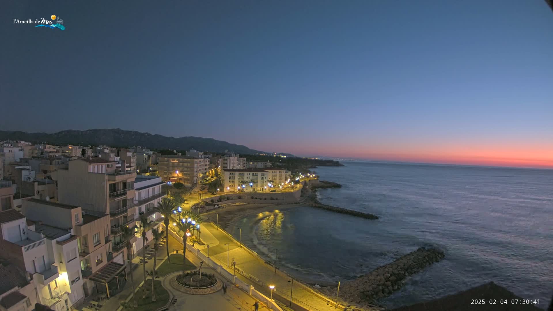 A coastal town at twilight, illuminated by streetlights, overlooks a calm sea under a mostly clear sky with a hint of pink on the horizon.