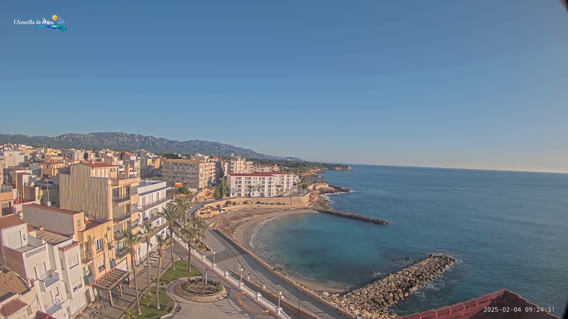 A coastal town with a small, crescent-shaped beach and calm, blue water under a clear, sunny sky and backed by mountains in the distance.
