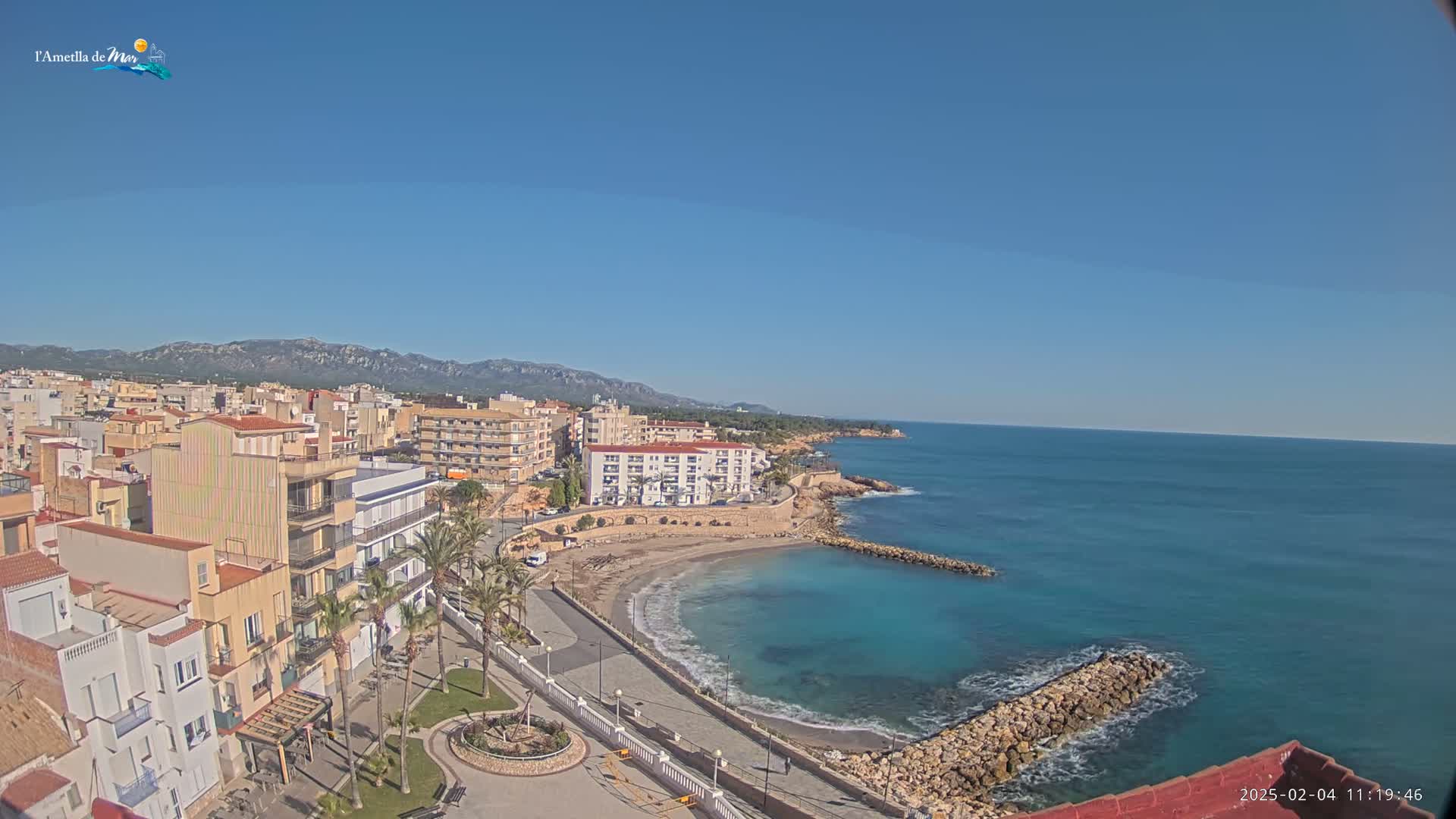 A coastal town with light-colored buildings, palm trees, and a small sandy beach is nestled against a calm turquoise sea under a clear blue sky.