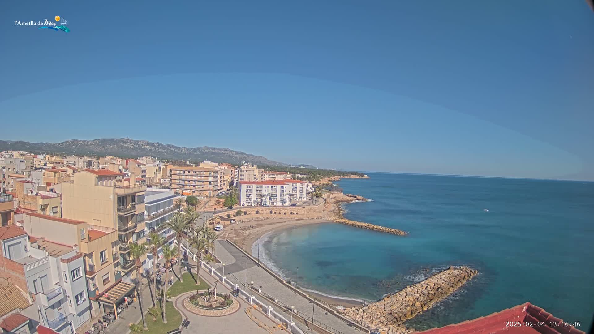 A coastal town with buildings and a small beach is seen on a clear, sunny day next to a calm, turquoise ocean and mountains in the distance.