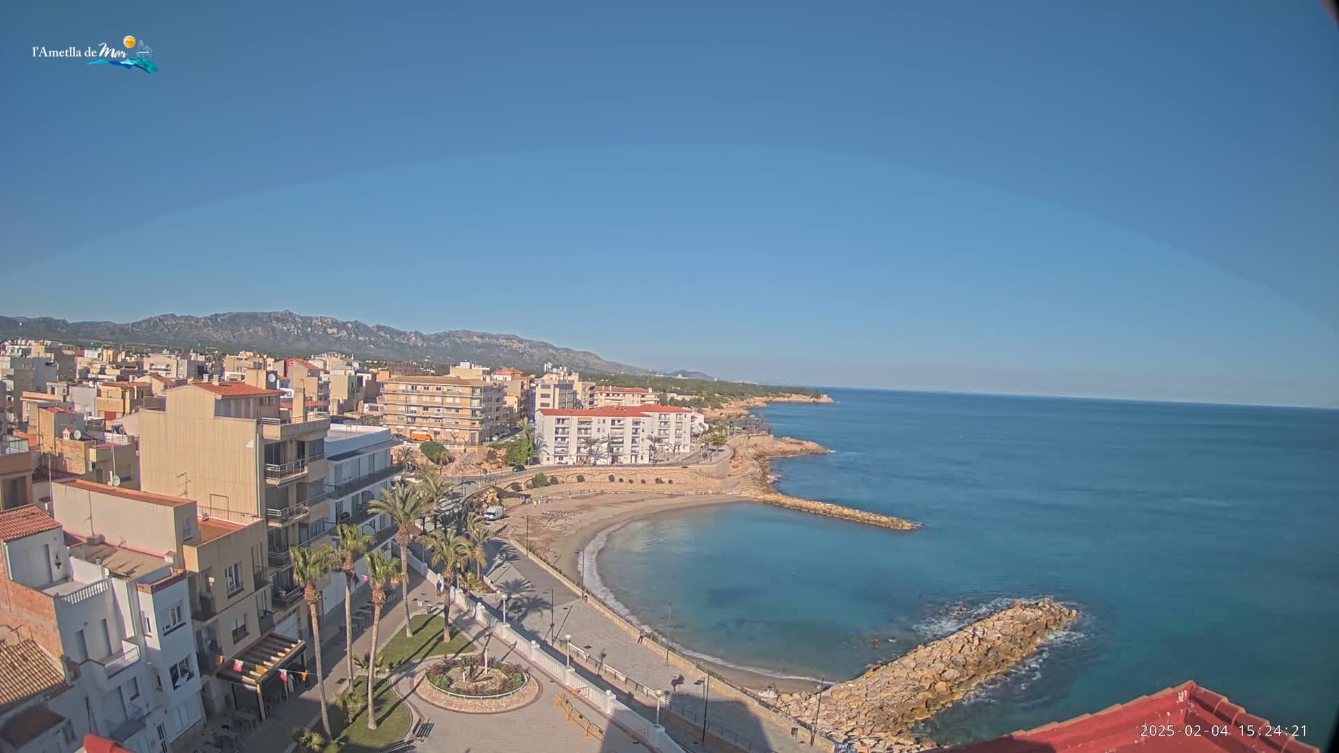 A coastal town with buildings, palm trees, and a small sandy beach curving into a calm blue sea under a clear blue sky.