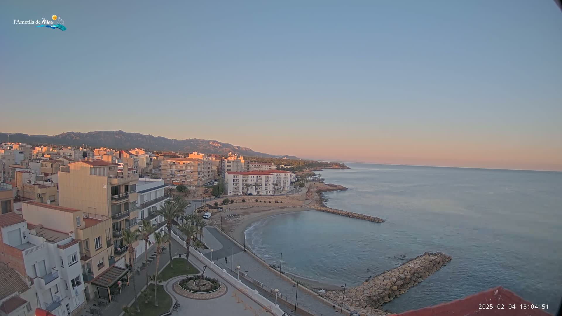 A coastal town with buildings and a small beach is seen at sunset under a clear sky, with calm water and mountains in the background.