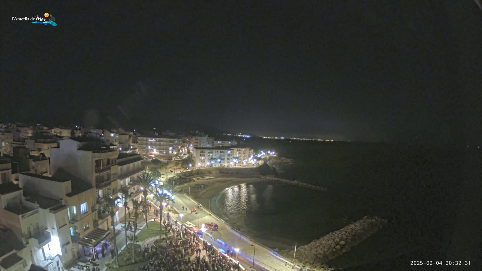 A nighttime aerial view shows a coastal town with a crowd gathered near the water, which is calm under a clear, dark sky.
