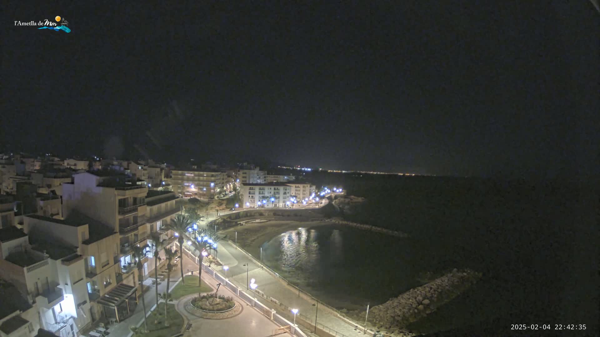 A nighttime aerial view shows a coastal town with buildings lit up along a calm, dark bay under a clear, dark sky.