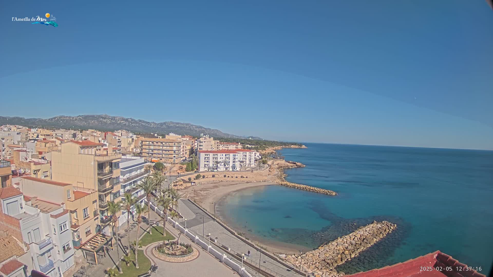 A coastal town with buildings, palm trees, and a small sandy beach is seen on a clear, sunny day with calm, turquoise water.