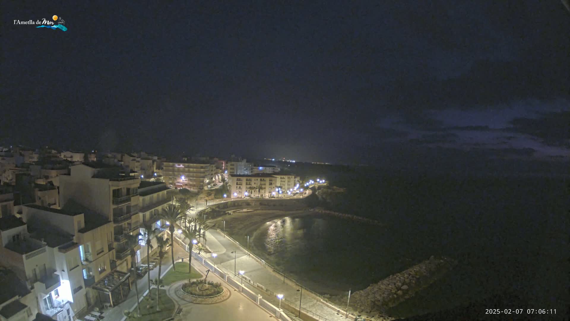 A nighttime, aerial view shows a coastal town with buildings illuminated along a calm bay under a dark sky.