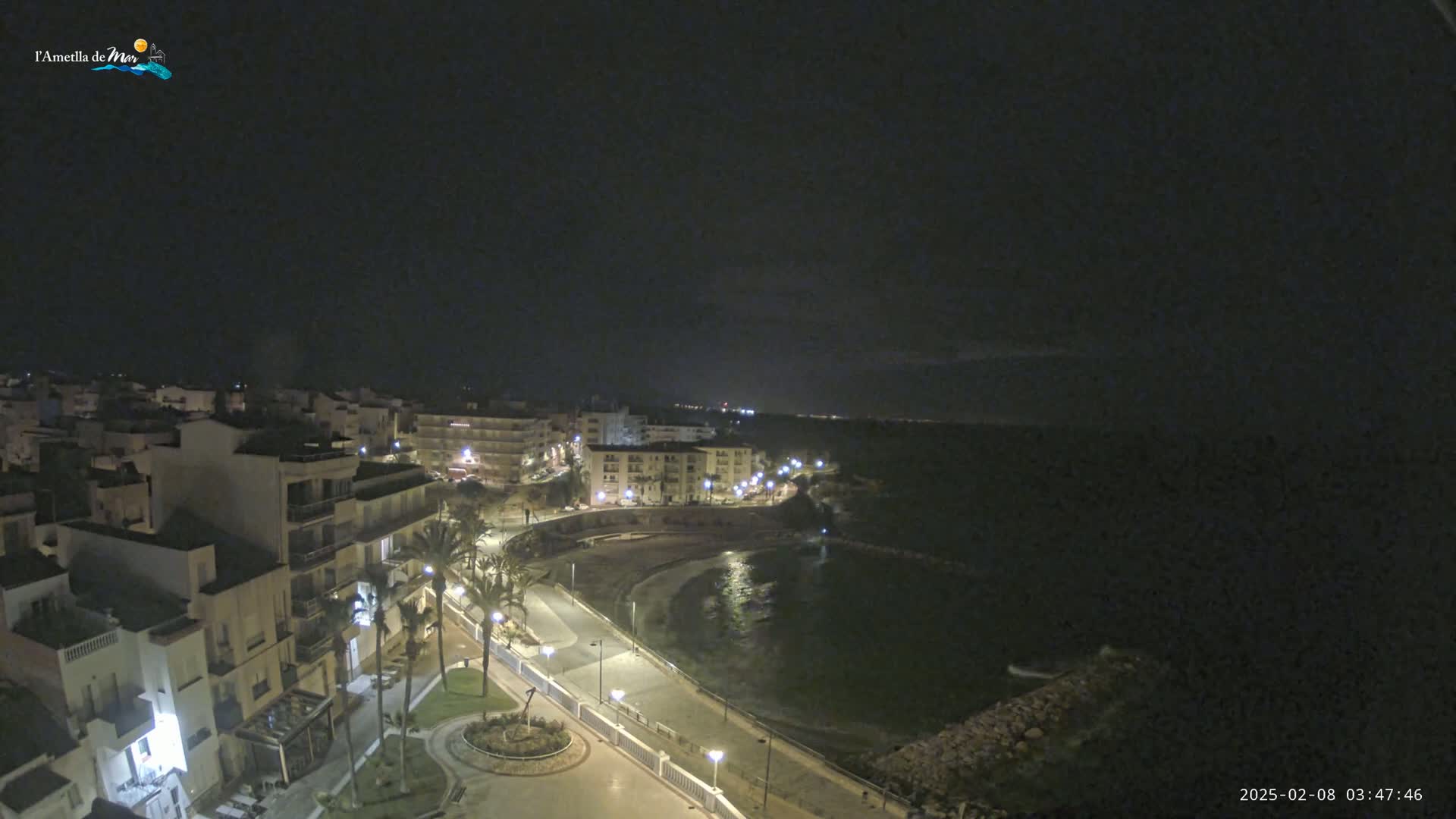 A nighttime coastal town scene shows buildings lit along a curved roadway next to a calm, dark bay under a clear, dark sky.