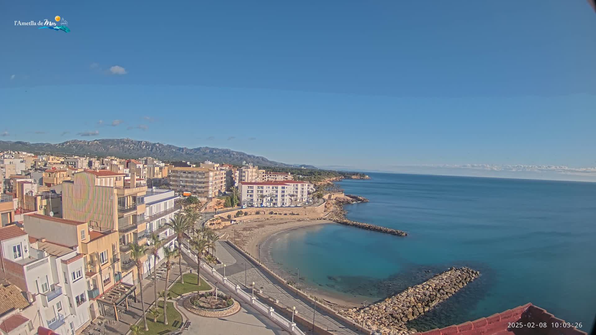 A coastal town with buildings and palm trees sits on a small, sandy beach next to a calm, blue sea under a clear, sunny sky, with mountains visible in the distance.