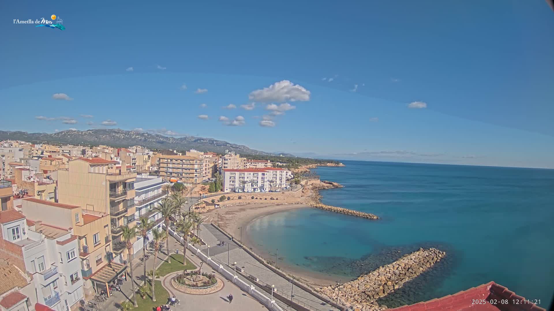 A coastal town with buildings, palm trees, and a small, curved beach sits against a clear blue sky with a few fluffy clouds, overlooking a calm, turquoise sea.