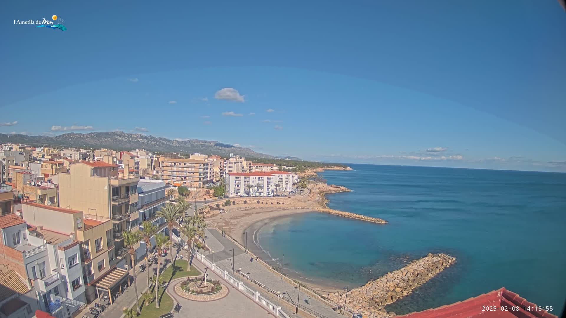 A coastal town with buildings and a small sandy cove nestled between rocky jetties, under a mostly clear blue sky with a few scattered clouds.