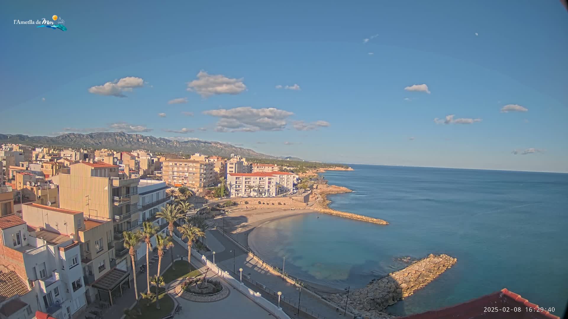 A coastal town with buildings, palm trees, and a small sandy beach sits under a mostly sunny sky with a few scattered clouds, overlooking a calm sea with rocky breakwaters.