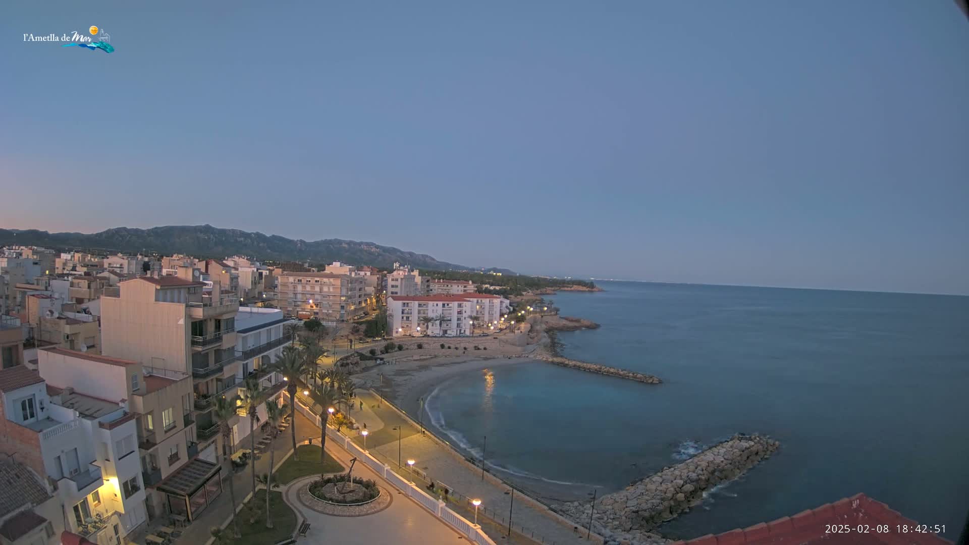 A coastal town at dusk is nestled against a mountain range, with a calm sea extending to the horizon under a clear sky.
