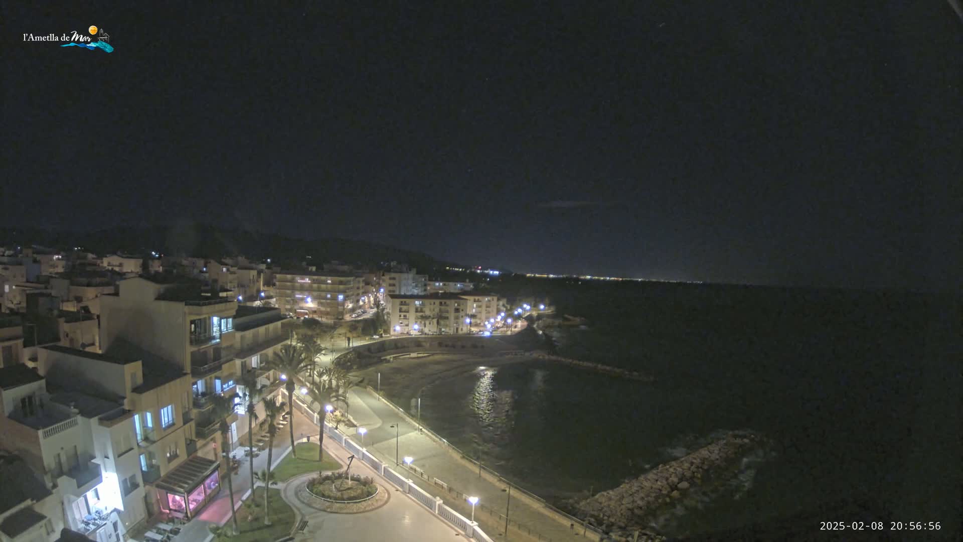 A nighttime, coastal town is illuminated, with a calm body of water visible along a paved walkway.